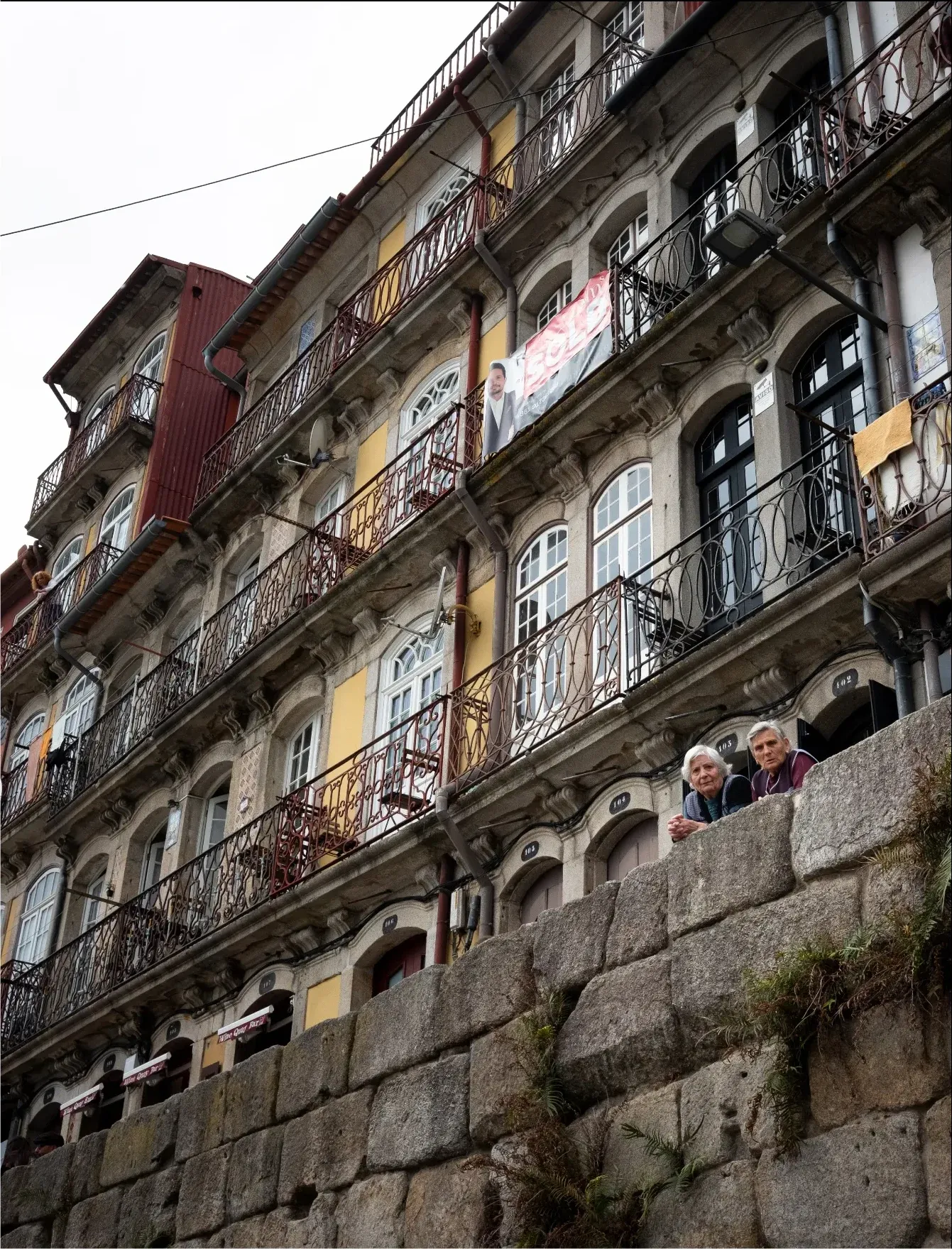 Dos señoras asomadas en un muro de piedra bajo edificios con balcones